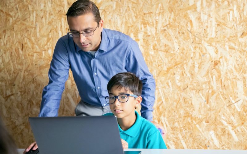 Middle-aged content teacher helping to boy with lesson and explaining theme. Young pupil in glasses looking at laptop computer screen and listening man. Childhood and digital education concept
