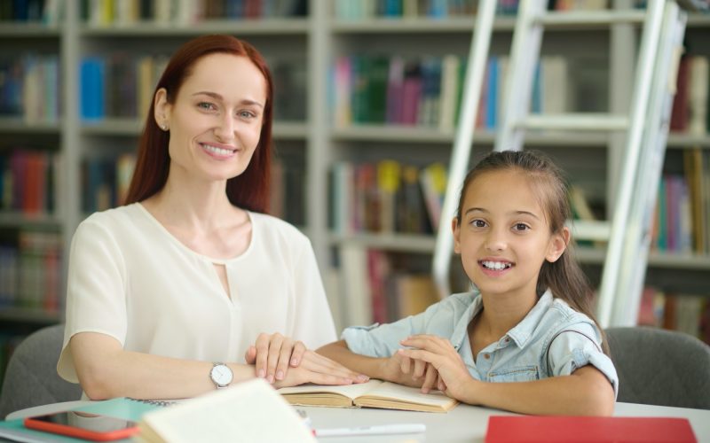Good mood. Smiling young long-haired woman and primary school girl in casual light clothing doing lessons at table in library looking at camera